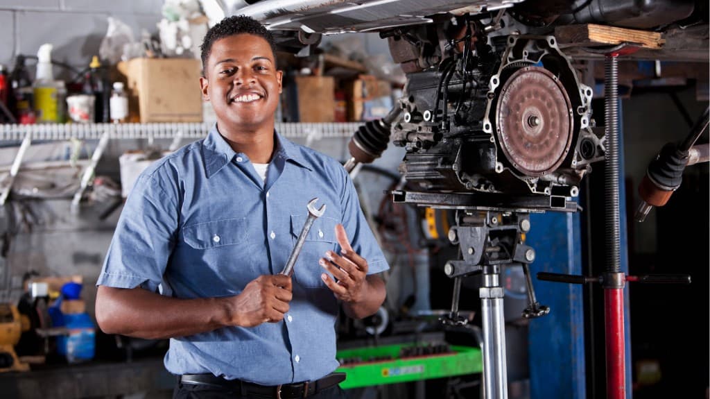 Smiling mechanic holding a wrench beside a transmission on a stand